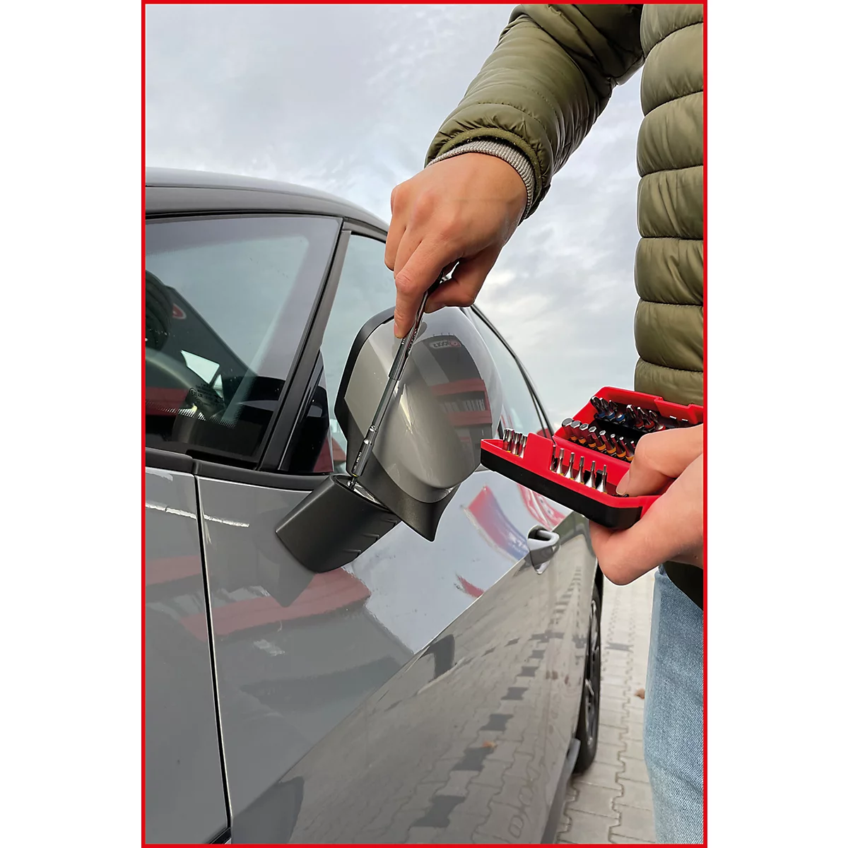 Person montiert Seitenspiegel an einem grauen Auto. In der Hand ein roter Werkzeugkasten mit Bits.